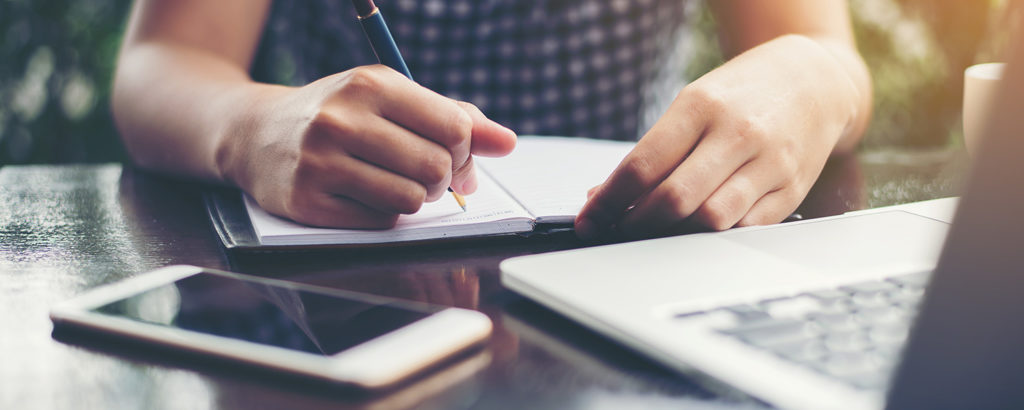 Man writing in a book with computer and cell phone in the foreground